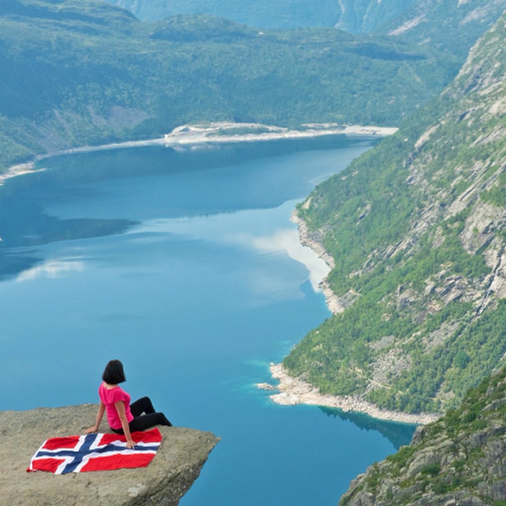 A woman sitting on a cliff overlooking a lake with a norwegian flag.