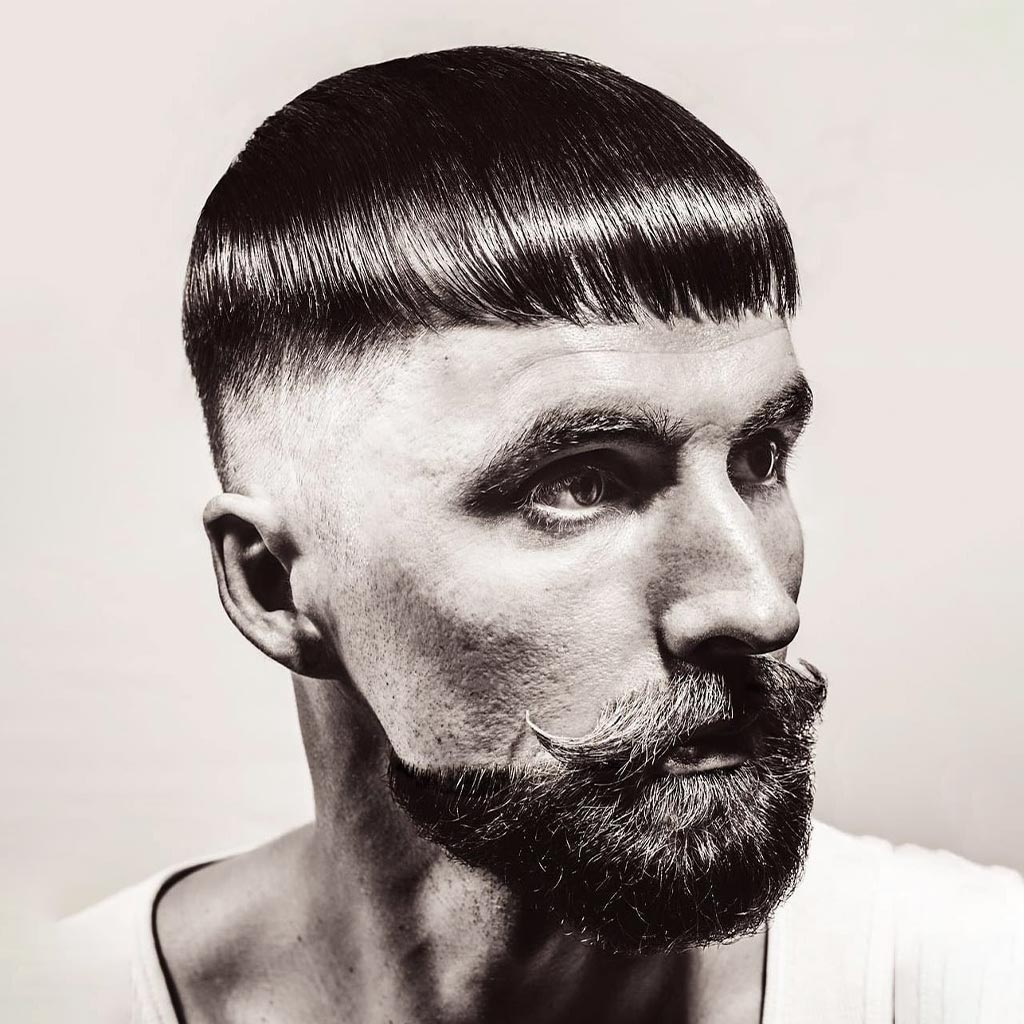 Black and white image of a person with a neatly styled, forward-combed short hairstyle featuring a smooth, rounded fringe. The person has a thick, styled beard and mustache, wearing a white top. The plain background enhances the Nordic elegance reminiscent of Scandinavia's timeless style.