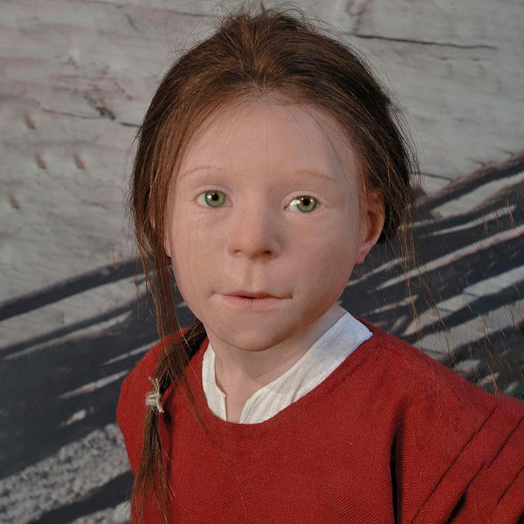 A realistic wax figure of a young Scandinavian girl with long brown hair in a braid, wearing a red garment. The background features a blurred image resembling a stone wall or natural setting.