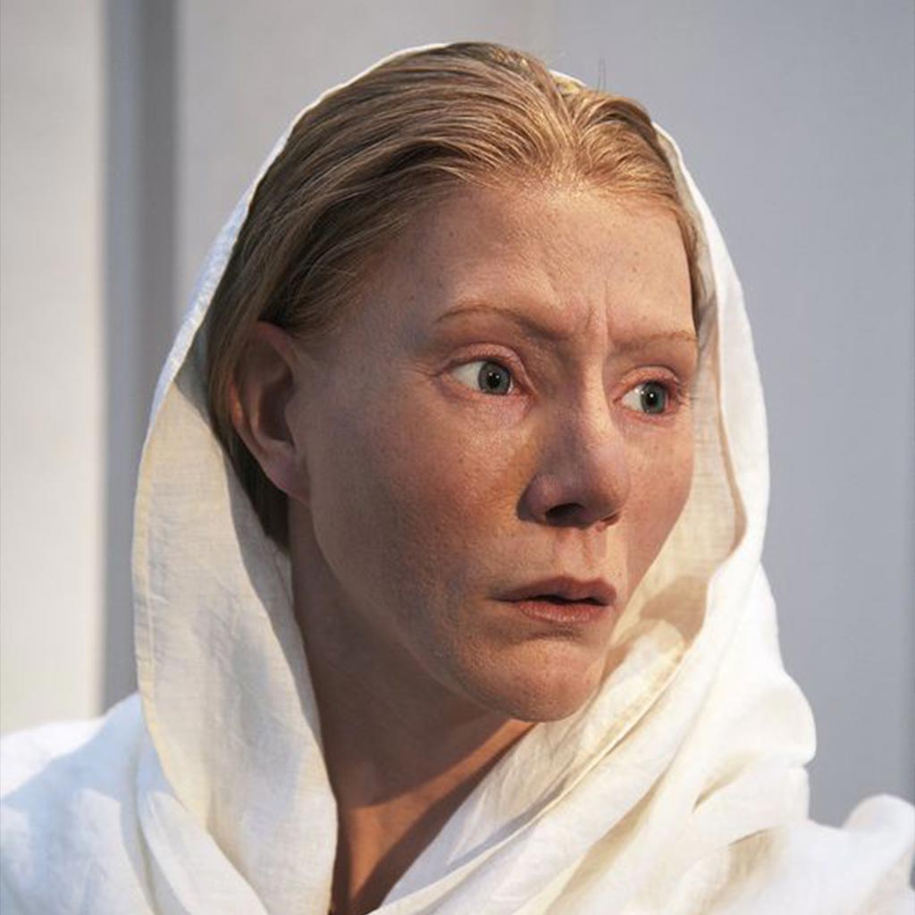 A realistic wax figure of a person with light skin is wearing a white hooded garment, evoking the serene yet enigmatic aura of Sweden. The expression is serious, with wide eyes and closed lips. The background is plain and softly lit.