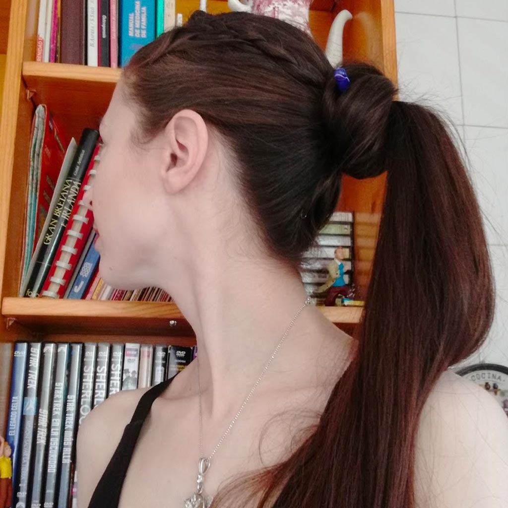 A person with long brown hair in a ponytail is turned to the side, facing a wooden bookshelf filled with books and DVDs. The person, exuding a Nordic elegance, is wearing a black top and a silver necklace.