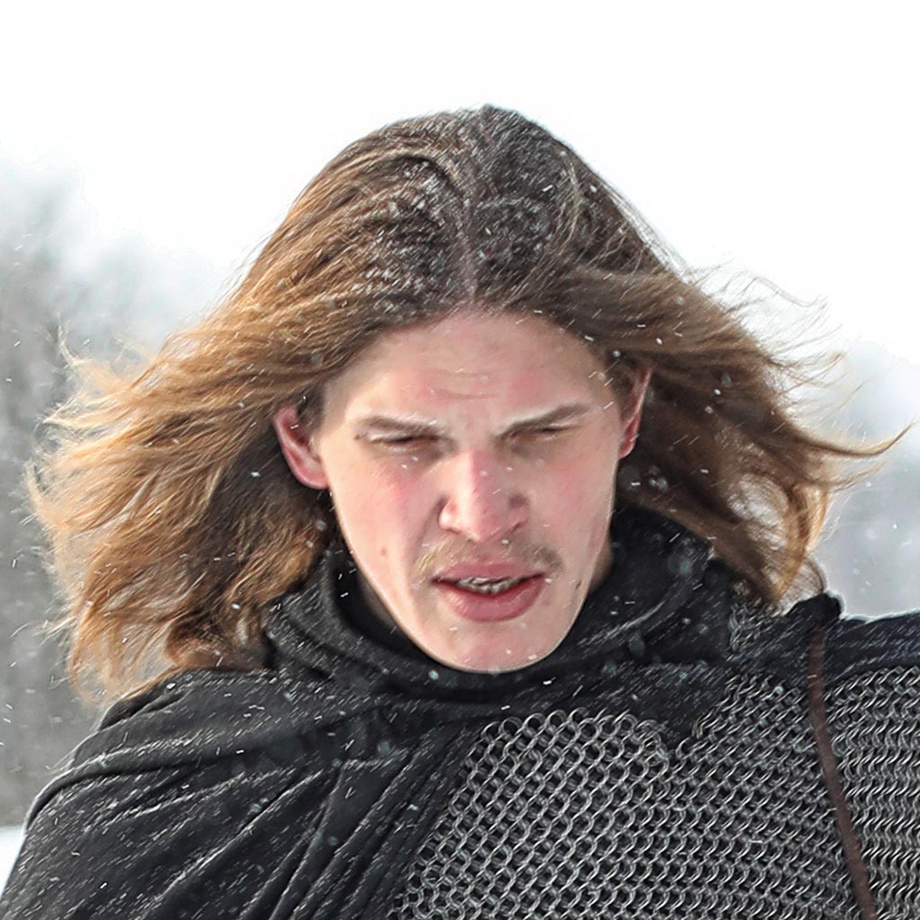 Amidst the snow, a person with long hair stands, wrapped in a chainmail garment and dark cloak. Their serious expression echoes the harsh beauty of Scandinavia, as snowflakes gently fall around them.