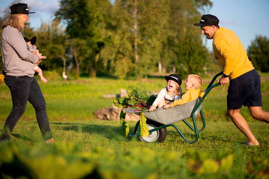 A man pushes a child in a wheelbarrow, both laughing, while another adult and a child watch with smiles, in a sunny, green outdoor setting.