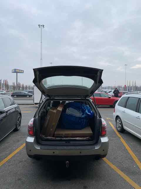 A silver car with its trunk open in a parking lot, revealing several large boxes and a blue bag inside. Other parked cars and a cloudy sky are visible in the background.
