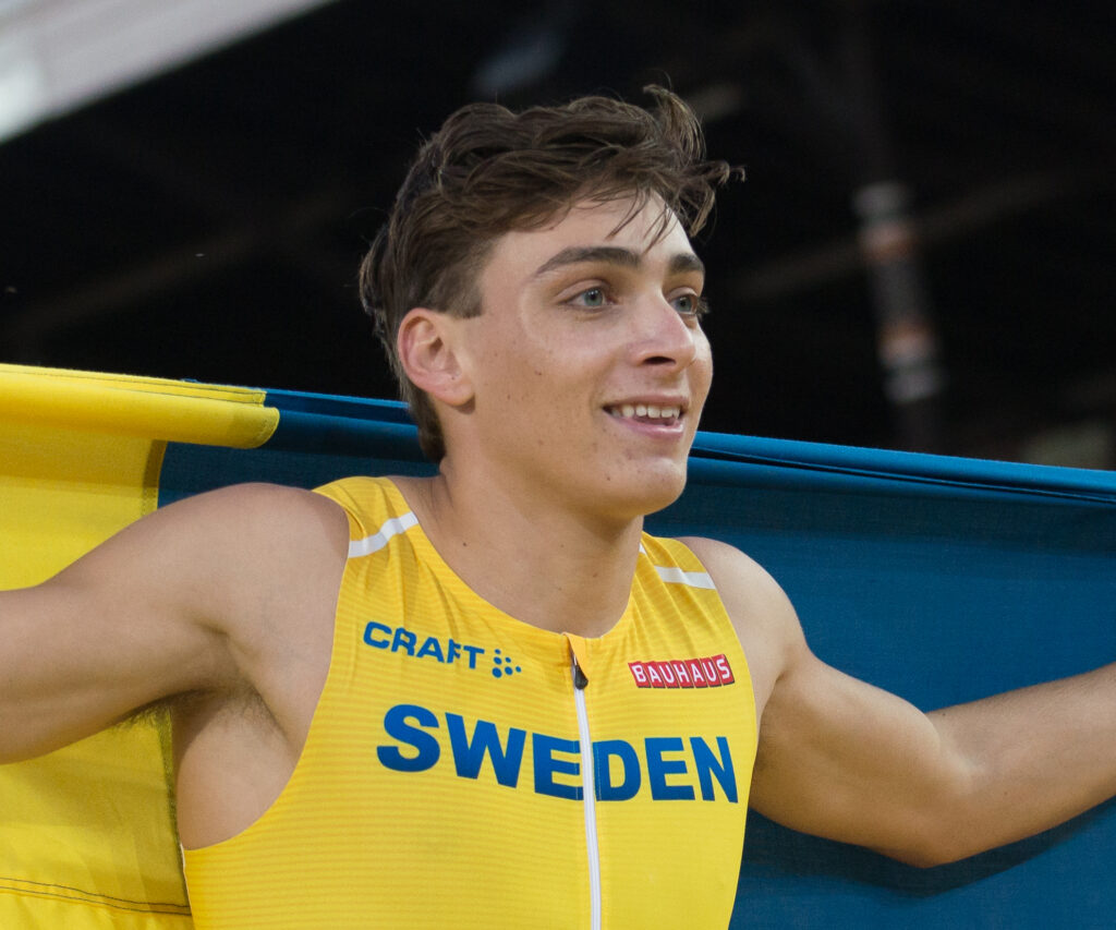 A male athlete - Armand Duplantis - wearing a yellow Sweden jersey holds a Swedish flag behind him. He appears to be celebrating, with a slight smile on his face. The background is blurred and dark.