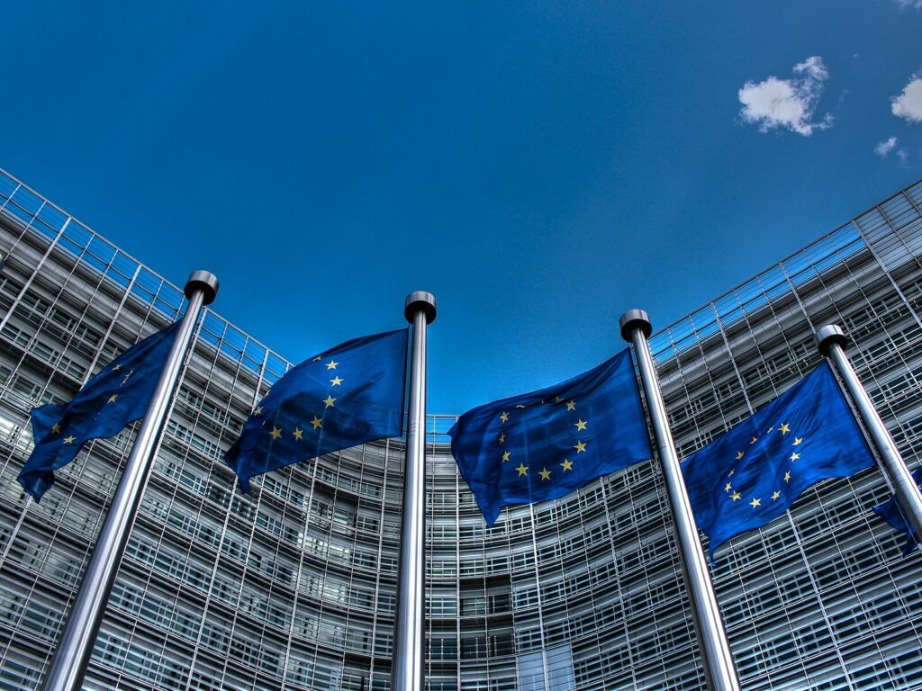 Four European Union flags on flagpoles wave in front of a modern glass building under a clear blue sky with a few clouds.