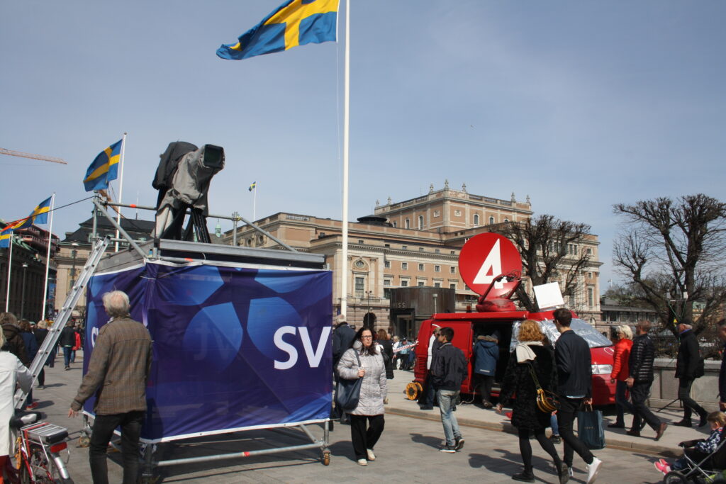 A group of people stand and walk in a city square near TV broadcast vans from SVT and TV4, with Swedish flags flying and a large building in the background under a clear sky.
