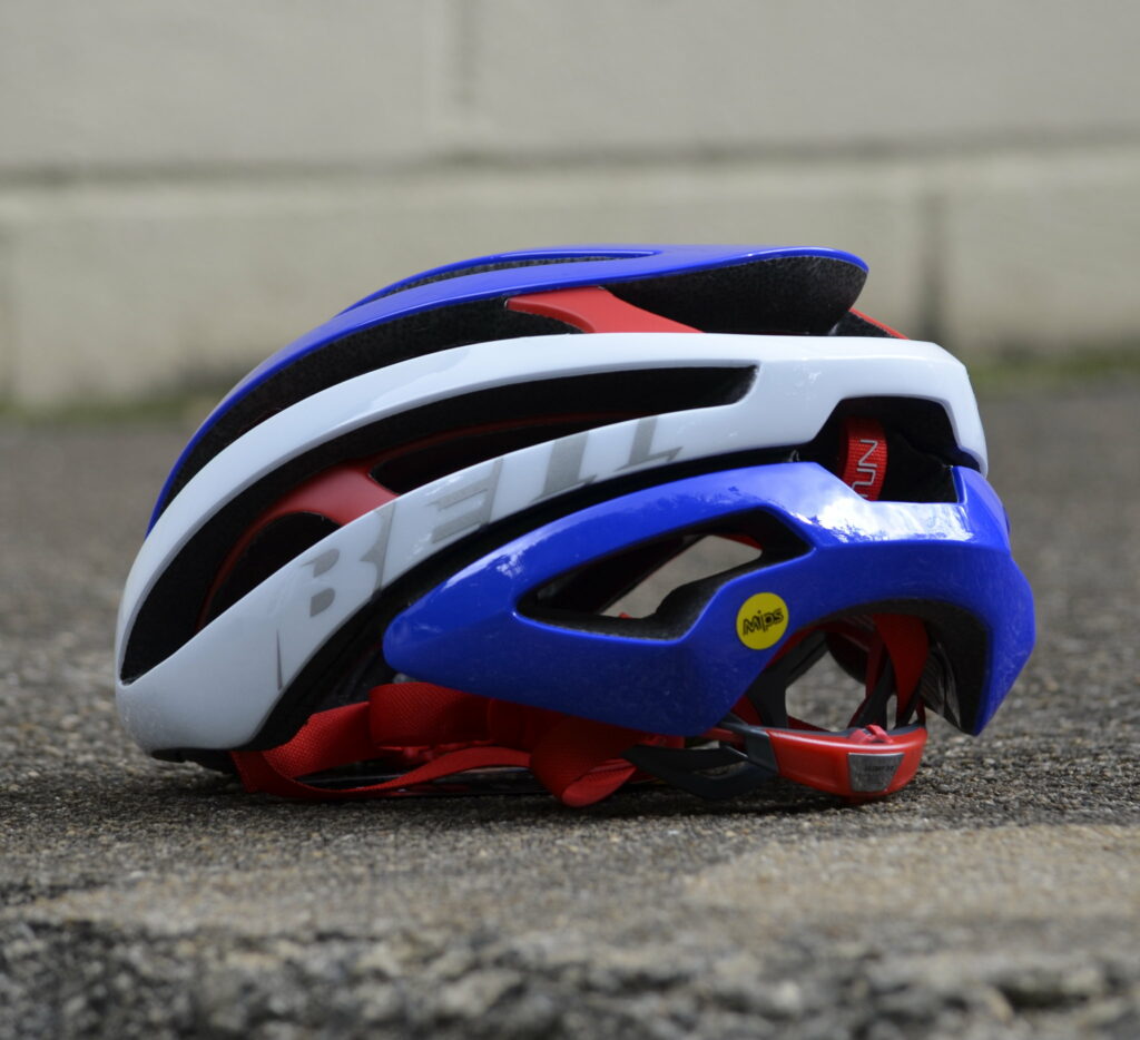A red, white, and blue bell cycling helmet is placed on the ground with a concrete wall in the background.