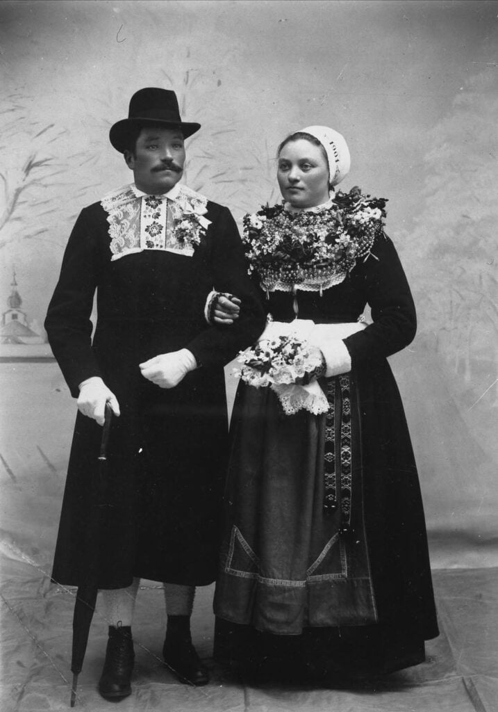 A man and woman in traditional folk costumes pose for a formal portrait. The man holds a cane and wears a hat, while the woman holds a bouquet and has a head covering. Both outfits feature intricate lace and embroidery details.